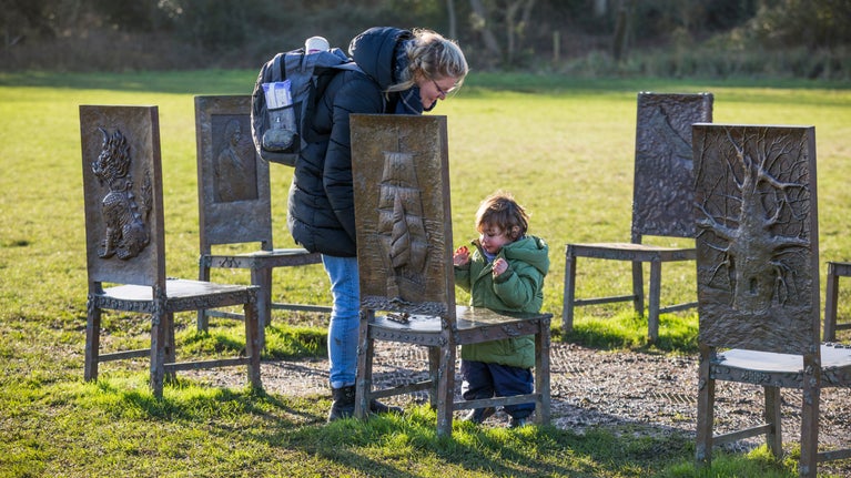 A mother and toddler at The Jurors artwork, a set of 12 bronze chairs in a field, at Runnymede, Surrey